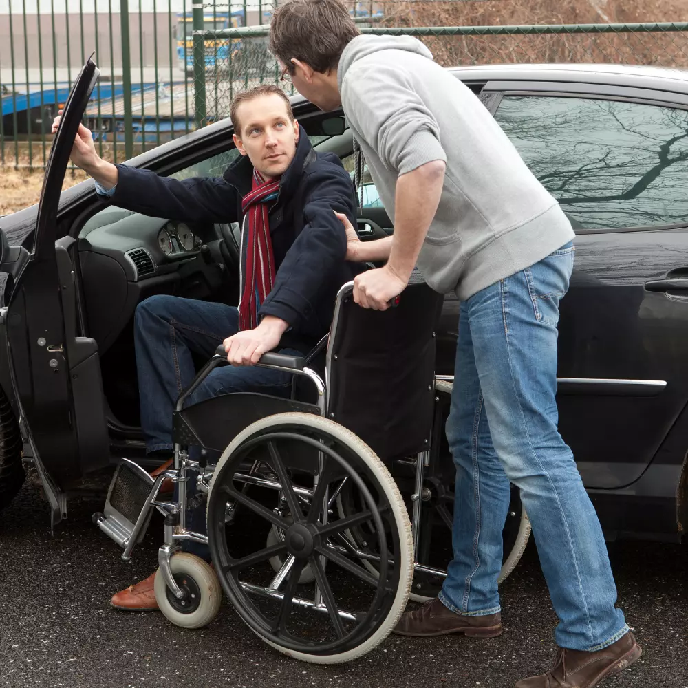 Man in wheelchair with carer helping him to get into car